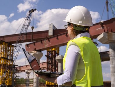 Female foreman working on laptop at bridge construction site. Girl builder controls erection of car road.