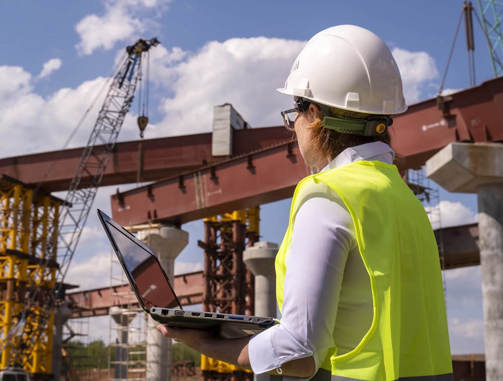 Female foreman working on laptop at bridge construction site. Girl builder controls erection of car road.